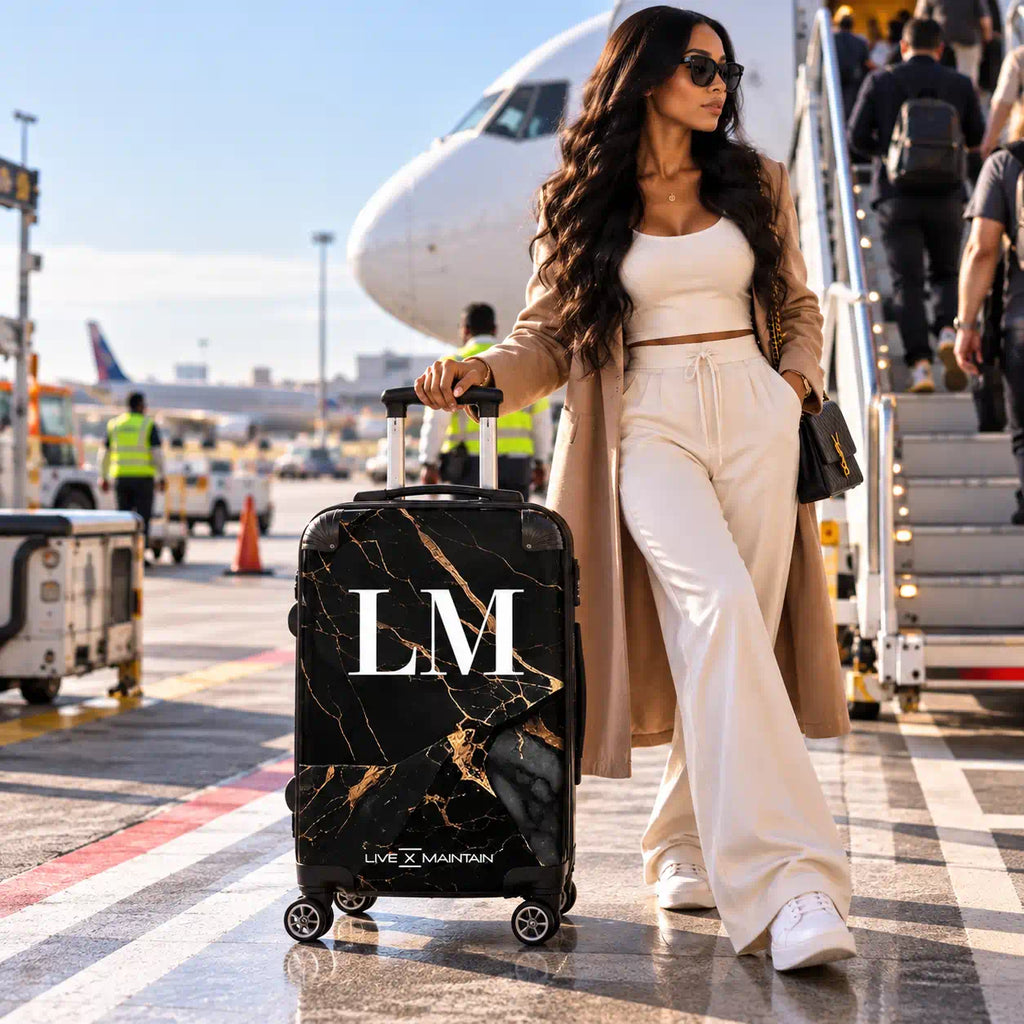 A woman in a beige trench coat and white outfit stands on an airport tarmac next to a black marble-patterned rolling suitcase with gold veining and large white 'LM' initials. An airplane and boarding stairs are visible in the background.