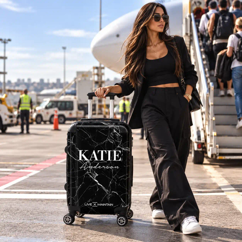 A woman in a black outfit stands on an airport tarmac next to a rolling suitcase with a black marble pattern. The suitcase has the name 'KATIE Anderson' printed on it in white script, and 'LIVE x MAINTAIN' is visible near the bottom.