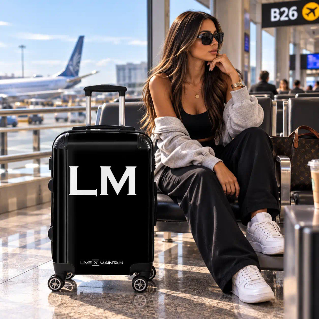 A woman sits in an airport lounge next to a black hard-shell suitcase with large white 'LM' initials and 'LIVE x MAINTAIN' branding. The scene includes an airplane, airport signage, and other travelers in the background.